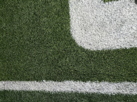 Close-up of a section of a football field showing green artificial turf with white painted lines. The texture of the grass and the distinct boundary markings are visible.