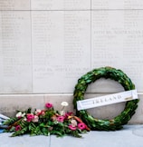 An engraved stone wall features a list of names, likely a memorial or monument. In front of it, a wreath made of green leaves is adorned with a white ribbon bearing the word 'Ireland'. A vibrant bouquet of pink and white flowers is placed beside the wreath, adding a touch of color and reverence to the solemn setting.