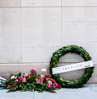 An engraved stone wall features a list of names, likely a memorial or monument. In front of it, a wreath made of green leaves is adorned with a white ribbon bearing the word 'Ireland'. A vibrant bouquet of pink and white flowers is placed beside the wreath, adding a touch of color and reverence to the solemn setting.