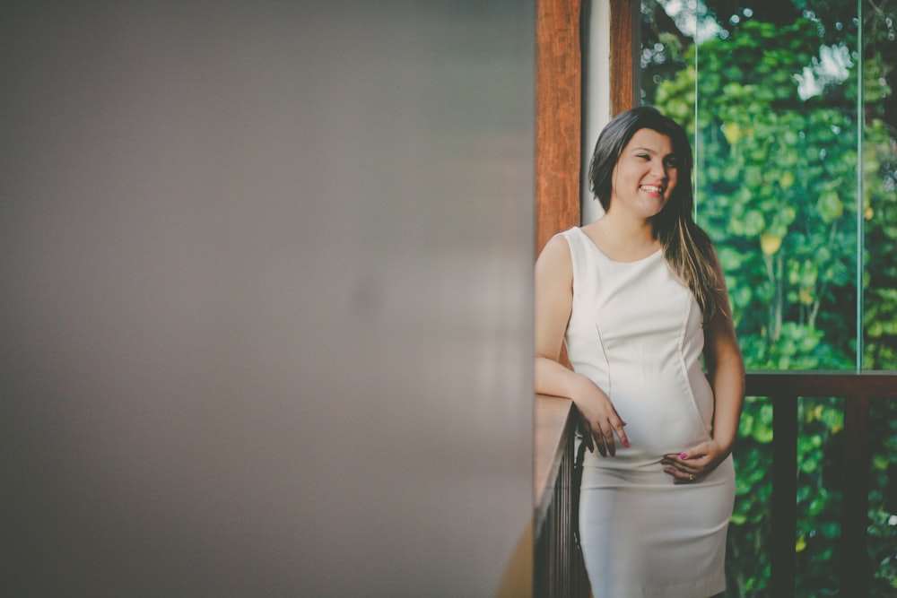 woman wearing white sleeveless dress leaning on brown wooden baluster inside room