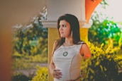 A woman in a white dress stands outside near a column, with lush greenery surrounding her. She appears thoughtful and slightly pensive, touching her abdomen gently. Sunlight filters through the foliage, creating a warm and serene atmosphere.