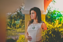 A woman in a white dress stands outside near a column, with lush greenery surrounding her. She appears thoughtful and slightly pensive, touching her abdomen gently. Sunlight filters through the foliage, creating a warm and serene atmosphere.