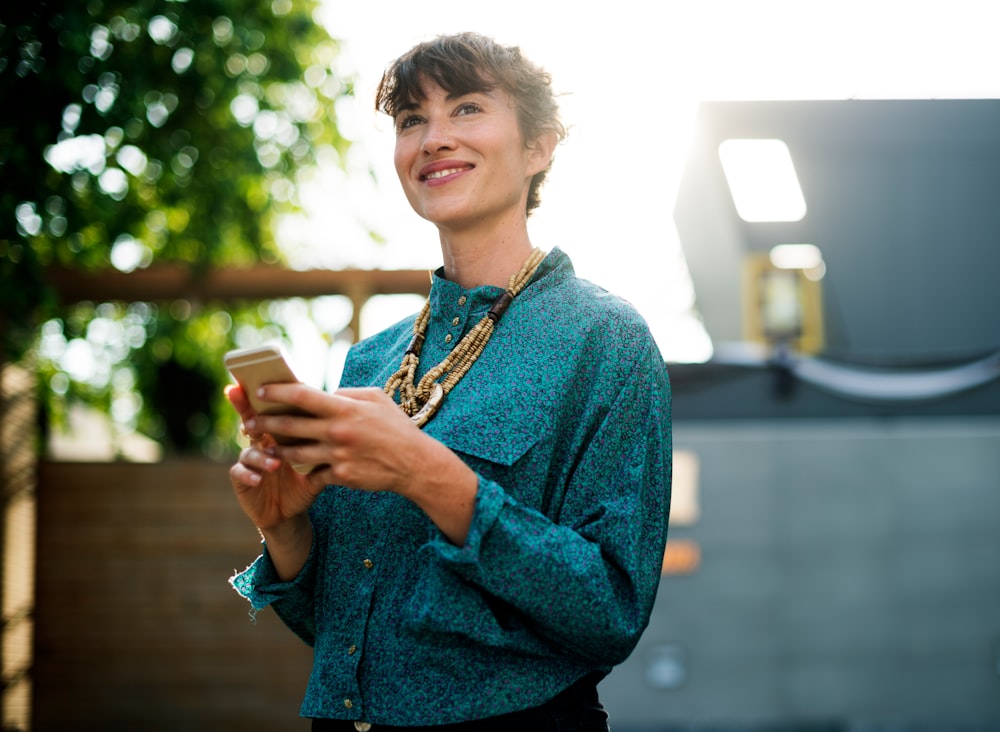 A smiling woman in an azure blouse holding a smartphone while standing outdoors
