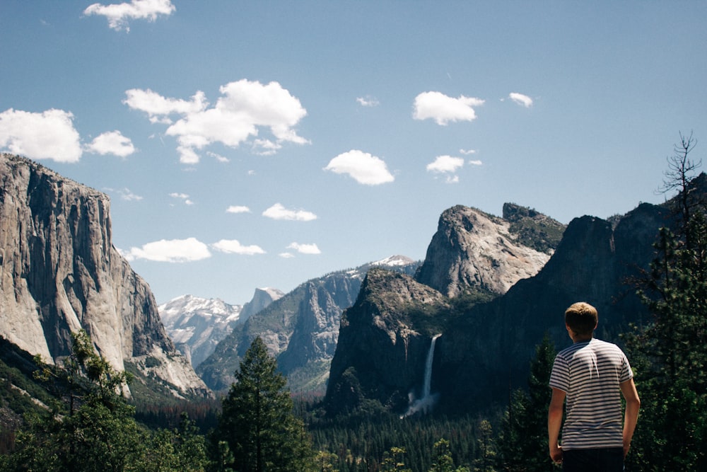 man standing next to mountains with waterfalls and trees