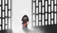 A figure resembling a Lego character stands in a space with glowing vertical lines on both sides. The character wears a dark cloak and hood, holding a bright red lightsaber. The background is filled with mist, creating a mysterious and dramatic atmosphere.