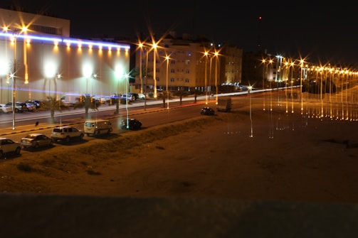 A nighttime urban scene with brightly illuminated buildings and streets. Streetlights emit a warm glow, creating starburst effects. Several cars are parked along the street, and light trails from moving vehicles suggest motion and activity.