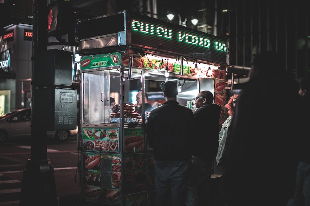 Dos hombres parados frente a un carrito de comida junto a un poste negro durante la noche