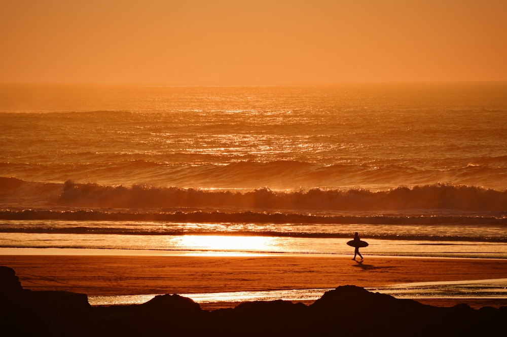 Persona sosteniendo una tabla de surf caminando por la orilla del mar durante la hora dorada