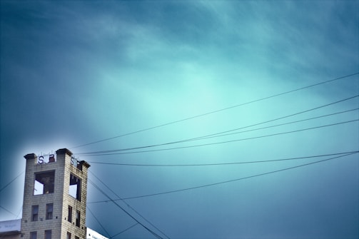 A tall, narrow building with a large S logo and an open top area is situated to the left. Overhead wires stretch across the sky, which is a vibrant gradient of blue. The building seems isolated against the expansive sky.