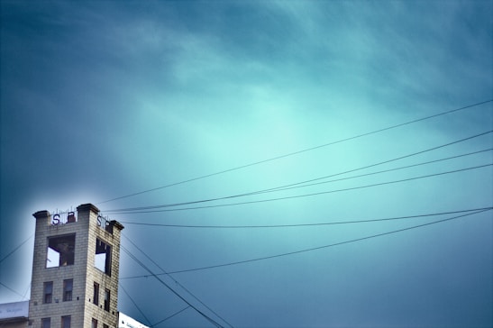 A tall, narrow building with a large S logo and an open top area is situated to the left. Overhead wires stretch across the sky, which is a vibrant gradient of blue. The building seems isolated against the expansive sky.
