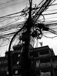 A complex tangle of electrical cables is wrapped around a utility pole, with a background of urban buildings featuring various signs and advertisements. The photograph is in high contrast black and white.