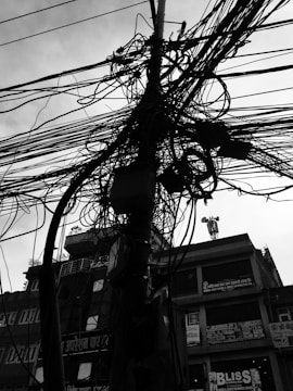 A complex tangle of electrical cables is wrapped around a utility pole, with a background of urban buildings featuring various signs and advertisements. The photograph is in high contrast black and white.