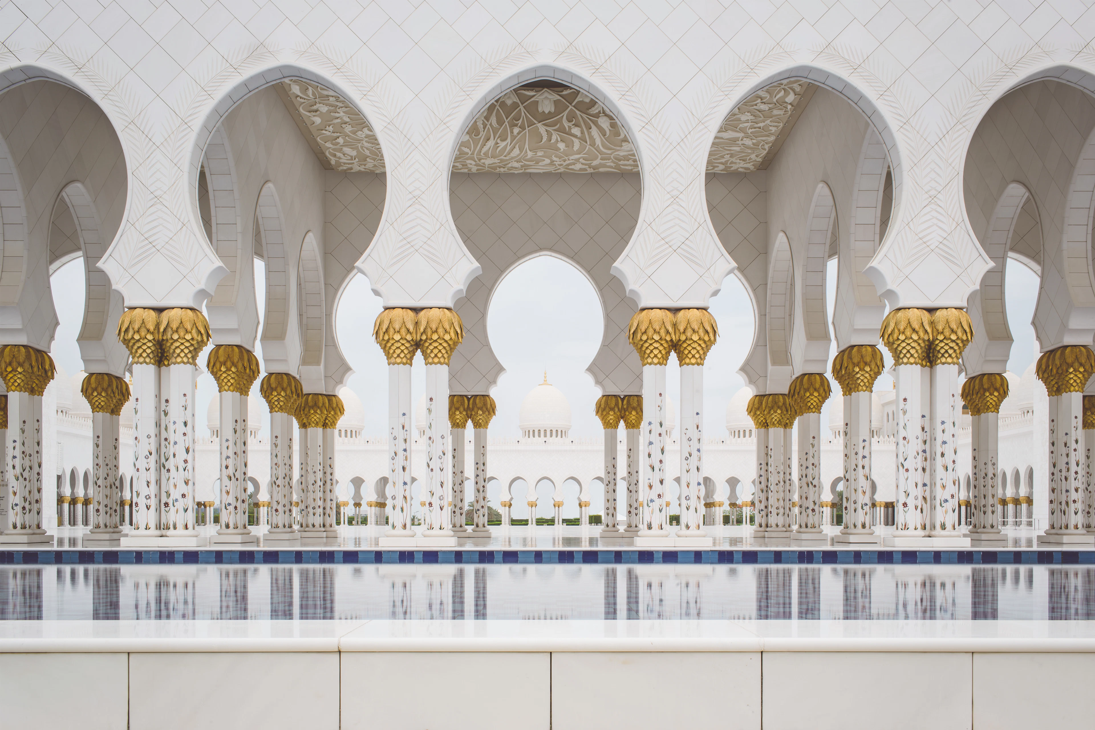 A white arcade with golden column capitals at the Sheikh Zayed Grand Mosque, reflected in water