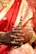 Intricate henna designs adorn the hands of a person wearing ornate gold bangles and a vibrant red saree. The fingers are clasped together, highlighting rich cultural symbolism. The saree features golden embroidery, adding to the luxurious appearance. A small, colorful wooden object is held delicately.