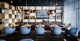 A modern restaurant interior features a row of light blue chairs arranged neatly in front of tables set with green napkins and condiments. Behind the seating area is a bookshelf with an assortment of decorative items, including books, small plants, and reflective spherical lamps. A chalkboard displays text inviting customers to try Irish coffee. The ambiance is enhanced by soft lighting and floor-to-ceiling sheer curtains that allow natural light to filter in.