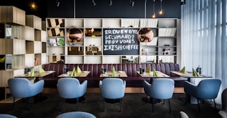 A modern restaurant interior features a row of light blue chairs arranged neatly in front of tables set with green napkins and condiments. Behind the seating area is a bookshelf with an assortment of decorative items, including books, small plants, and reflective spherical lamps. A chalkboard displays text inviting customers to try Irish coffee. The ambiance is enhanced by soft lighting and floor-to-ceiling sheer curtains that allow natural light to filter in.