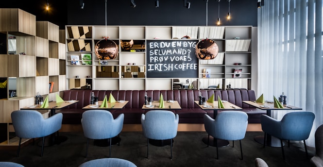 A modern restaurant interior features a row of light blue chairs arranged neatly in front of tables set with green napkins and condiments. Behind the seating area is a bookshelf with an assortment of decorative items, including books, small plants, and reflective spherical lamps. A chalkboard displays text inviting customers to try Irish coffee. The ambiance is enhanced by soft lighting and floor-to-ceiling sheer curtains that allow natural light to filter in.