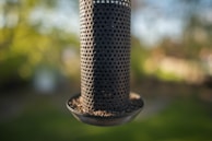 A cylindrical metal bird feeder with numerous small holes filled with seeds, set against a blurred green and yellow natural background.