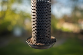 A cylindrical metal bird feeder with numerous small holes filled with seeds, set against a blurred green and yellow natural background.