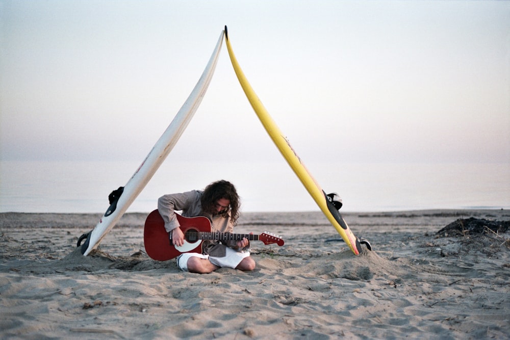 man playing guitar sitting on gray sand under cloudy sky