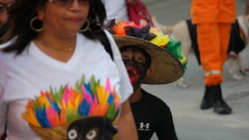 A person wearing a white t-shirt with a colorful print is walking alongside another individual in a black outfit with vibrant, multicolored feathers on their hat. The background shows a person in orange clothing, and a dog walking nearby.
