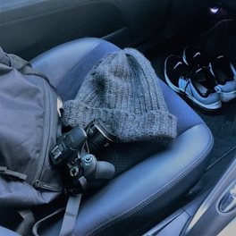 A gray knitted hat, a digital camera, and a backpack are placed on a dark car seat. A pair of black and white sneakers is on the car floor.