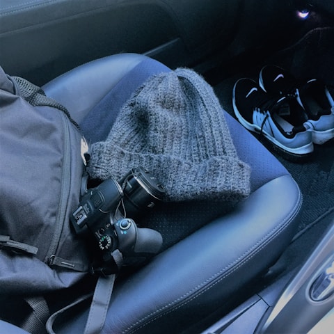 A gray knitted hat, a digital camera, and a backpack are placed on a dark car seat. A pair of black and white sneakers is on the car floor.