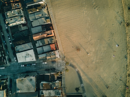 An aerial view of a cityscape with tightly packed buildings on one side and an open sandy area on the other. Vehicles are parked along the streets, and the shadow of streetlights can be seen on the sand. The transition from urban to open space is clearly visible.