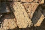 Large, rough-hewn stone slabs with an irregular arrangement. The surface displays a mix of warm earthy tones and is textured with visible striations and natural markings. Shadows are cast across the stones, adding depth to the composition.