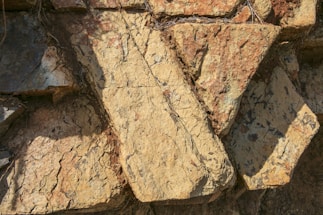 Large, rough-hewn stone slabs with an irregular arrangement. The surface displays a mix of warm earthy tones and is textured with visible striations and natural markings. Shadows are cast across the stones, adding depth to the composition.