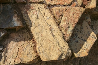 Large, rough-hewn stone slabs with an irregular arrangement. The surface displays a mix of warm earthy tones and is textured with visible striations and natural markings. Shadows are cast across the stones, adding depth to the composition.
