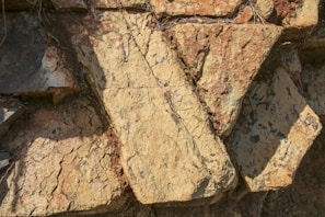 Large, rough-hewn stone slabs with an irregular arrangement. The surface displays a mix of warm earthy tones and is textured with visible striations and natural markings. Shadows are cast across the stones, adding depth to the composition.