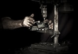 Hands operating a metal drill press in a dimly lit workshop. The focus is on the hands and the machine, highlighting the details of the tools and the worn, textured skin of the worker.
