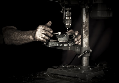 Hands operating a metal drill press in a dimly lit workshop. The focus is on the hands and the machine, highlighting the details of the tools and the worn, textured skin of the worker.