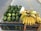 A street market display featuring a collection of large, green avocados arranged neatly on a cardboard-covered crate next to several bunches of ripe yellow bananas with brown spots. The scene is set on a paved sidewalk.