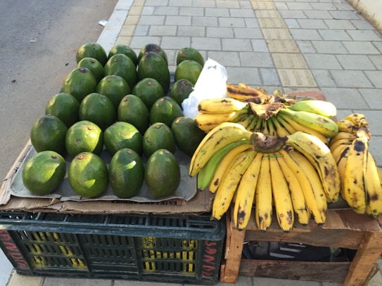 A street market display featuring a collection of large, green avocados arranged neatly on a cardboard-covered crate next to several bunches of ripe yellow bananas with brown spots. The scene is set on a paved sidewalk.