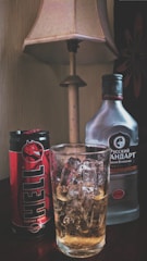 A can of Hell energy drink and a bottle of Russian Standard vodka are placed on a table next to a glass filled with ice cubes and a light-colored liquid. There is a table lamp with a beige shade in the background.