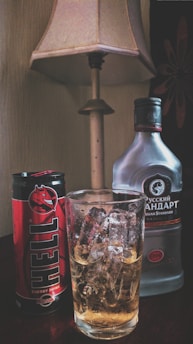A can of Hell energy drink and a bottle of Russian Standard vodka are placed on a table next to a glass filled with ice cubes and a light-colored liquid. There is a table lamp with a beige shade in the background.