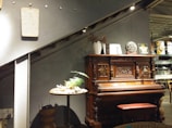 A vintage wooden piano with ornate carvings is positioned against a wall beneath a stairway. On the piano are decorative items, including a sculpted head and various small artworks. To the left, a round table holds a small plant in a shallow dish. The setting appears to be in an indoor area with a modern but cozy ambiance.
