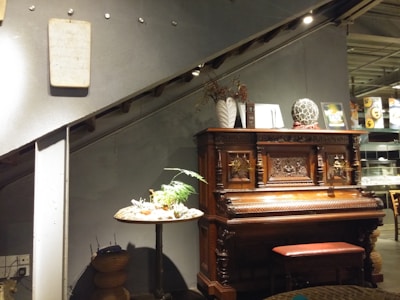 A vintage wooden piano with ornate carvings is positioned against a wall beneath a stairway. On the piano are decorative items, including a sculpted head and various small artworks. To the left, a round table holds a small plant in a shallow dish. The setting appears to be in an indoor area with a modern but cozy ambiance.
