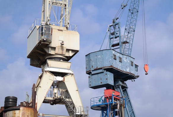 Two large industrial cranes are set against a clear blue sky. The left crane is mostly white with a rusted base, while the right crane is predominantly blue with visible wear and rust. Both cranes have complex structural designs with numerous wires and machinery. A red hoisting hook is visible on the right crane.