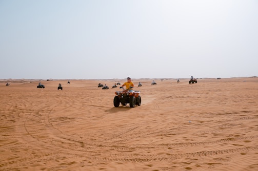 Numerous people are riding all-terrain vehicles (ATVs) on an expansive desert landscape. The desert is vast, with sand dunes stretching into the distance under a clear blue sky. Tire tracks crisscross the sandy surface, indicating the dynamic movement across the terrain. The scene conveys a sense of adventure and excitement.