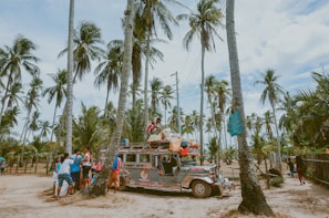 A colorful jeepney is parked on a sandy path surrounded by tall palm trees. Several people are gathered around the vehicle, some wearing brightly colored clothing. The scene appears tropical and vibrant, conveying a sense of community and travel.