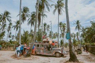 A colorful jeepney is parked on a sandy path surrounded by tall palm trees. Several people are gathered around the vehicle, some wearing brightly colored clothing. The scene appears tropical and vibrant, conveying a sense of community and travel.