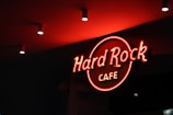 A neon sign displaying the words 'Hard Rock Cafe' in bright red is illuminated against a dark background. The ceiling is adorned with recessed lighting.