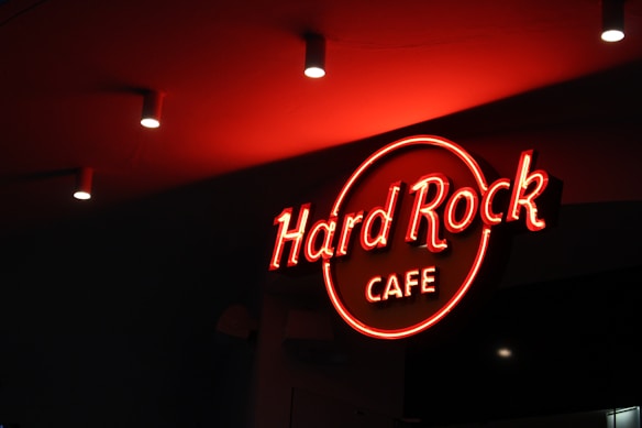 A neon sign displaying the words 'Hard Rock Cafe' in bright red is illuminated against a dark background. The ceiling is adorned with recessed lighting.