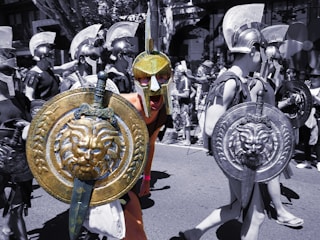 A group of people dressed in Roman or Greek warrior costumes participate in a parade or event. They are wearing helmets and carrying round shields with intricate designs, including lion motifs. The image has a mix of color and black-and-white elements, creating a striking contrast.