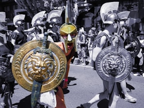A group of people dressed in Roman or Greek warrior costumes participate in a parade or event. They are wearing helmets and carrying round shields with intricate designs, including lion motifs. The image has a mix of color and black-and-white elements, creating a striking contrast.