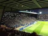 A large crowd in a covered stadium is illuminated by numerous phones with flashlights held by spectators. The audience is densely packed in the stands, while a brightly lit soccer field is visible on the right with a few players on it.