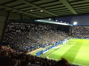 A large crowd in a covered stadium is illuminated by numerous phones with flashlights held by spectators. The audience is densely packed in the stands, while a brightly lit soccer field is visible on the right with a few players on it.
